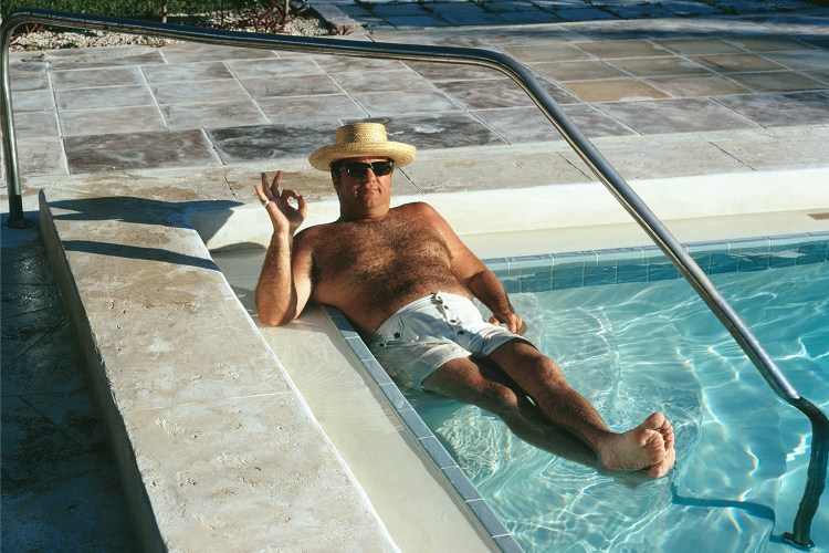 A dad in a straw hat with sunglasses and white swim trunks sitting in the corner of a pool making the OK sign