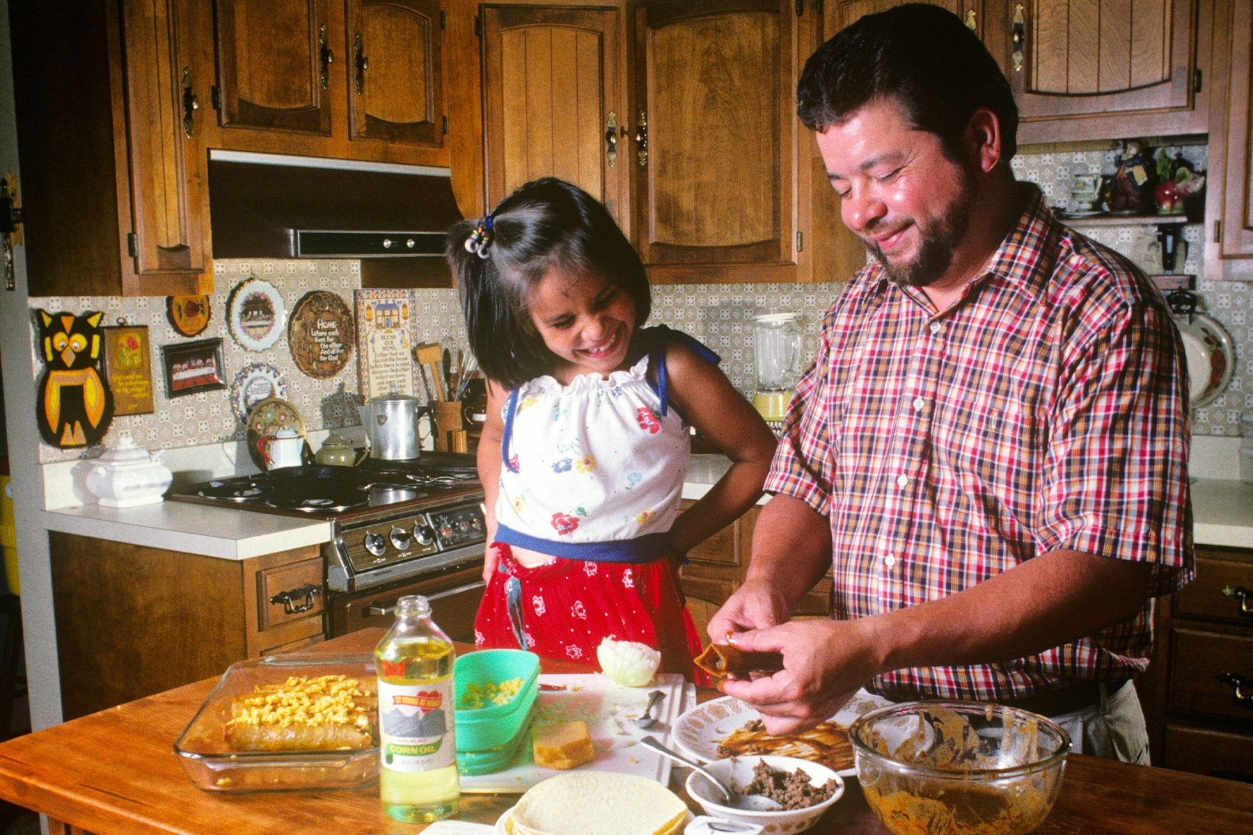 1980s Smiling father and daughter preparing food on finished wooden kitchen counter.