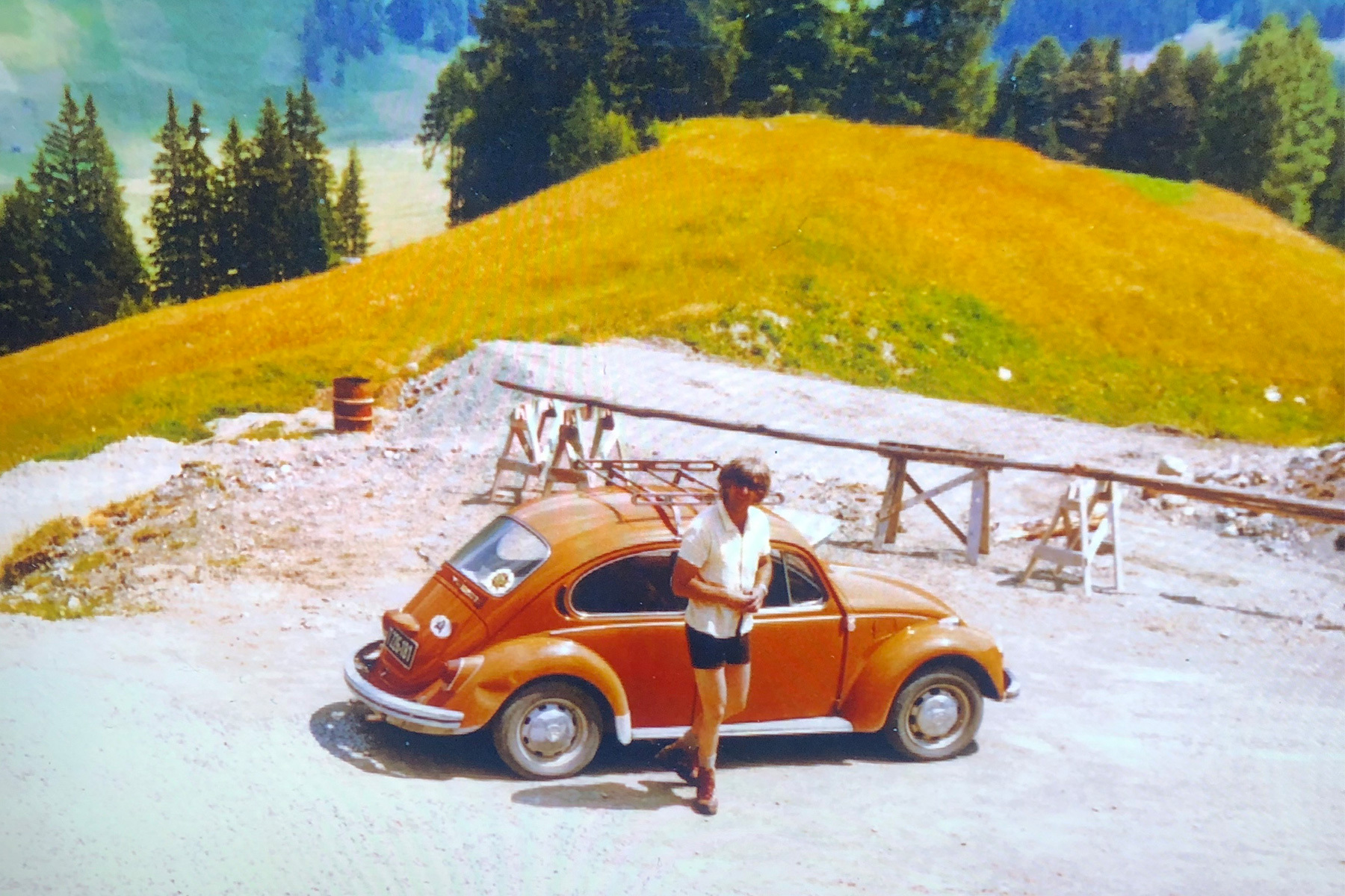 A dad in a short-sleeve button-up and shorts stands in front of an orange Volkswagen Beetle in a bucolic setting