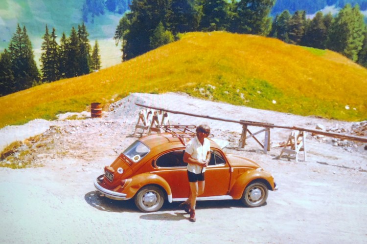 A dad in a short-sleeve button-up and shorts stands in front of an orange Volkswagen Beetle in a bucolic setting