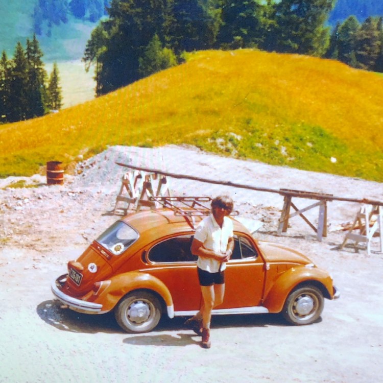 A dad in a short-sleeve button-up and shorts stands in front of an orange Volkswagen Beetle in a bucolic setting