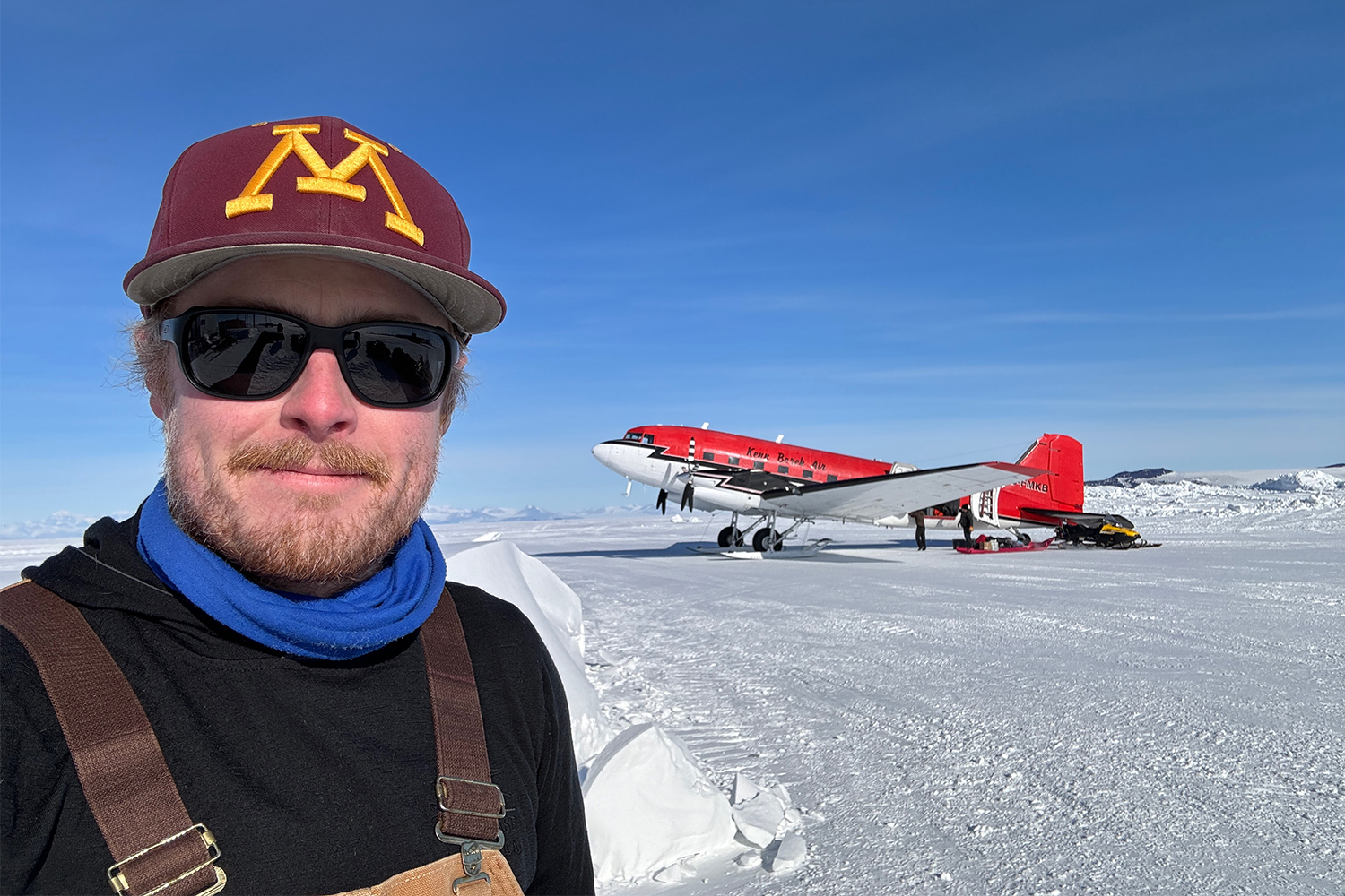 Dr. Peter Neff, a climate scientist, seen in black sunglasses and a maroon and gold hat with "M" on it with an airplane on ice in the background