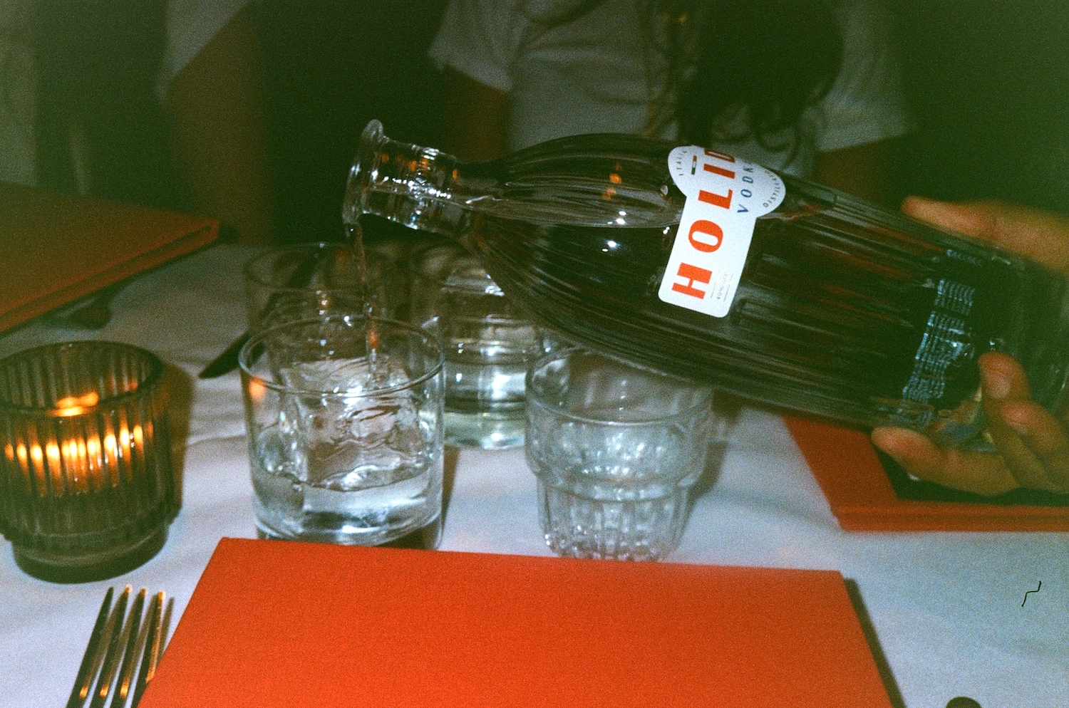 a hand pouring a bottle of holiday vodka into rocks glasses on a white table with red menus