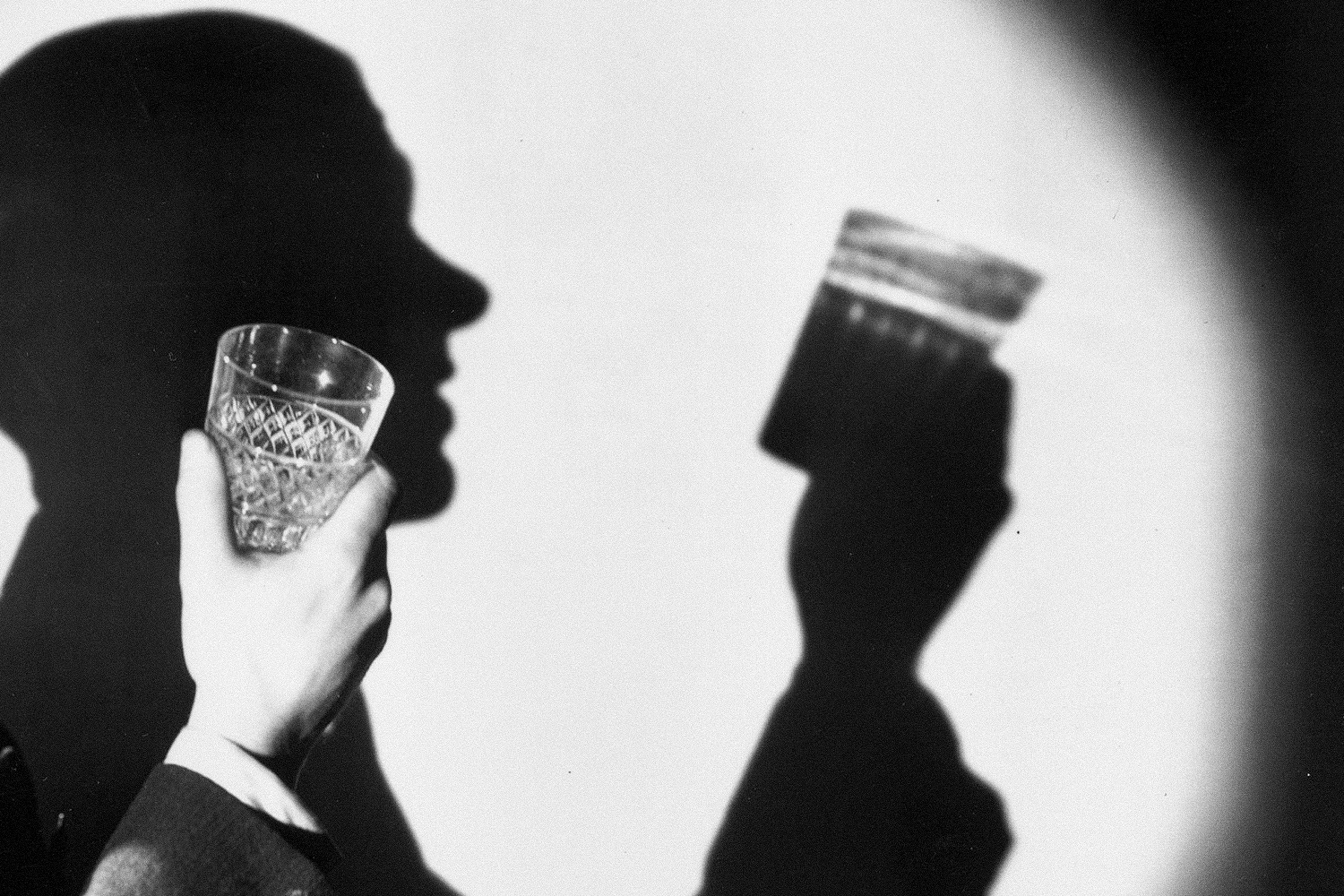 A semi-hidden man holding up a glass in a black and white picture. The photo emphasizes his shadow examining the glass.