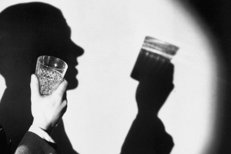 A semi-hidden man holding up a glass in a black and white picture. The photo emphasizes his shadow examining the glass.