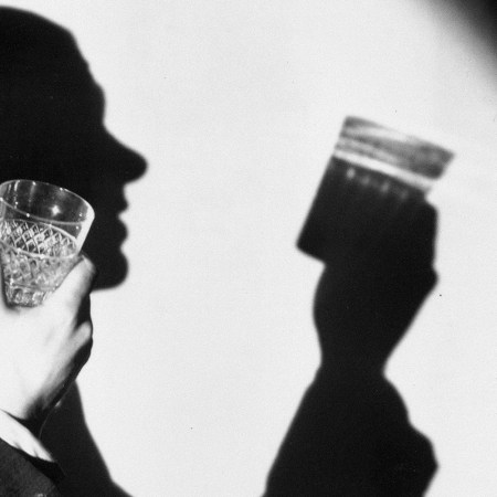 A semi-hidden man holding up a glass in a black and white picture. The photo emphasizes his shadow examining the glass.