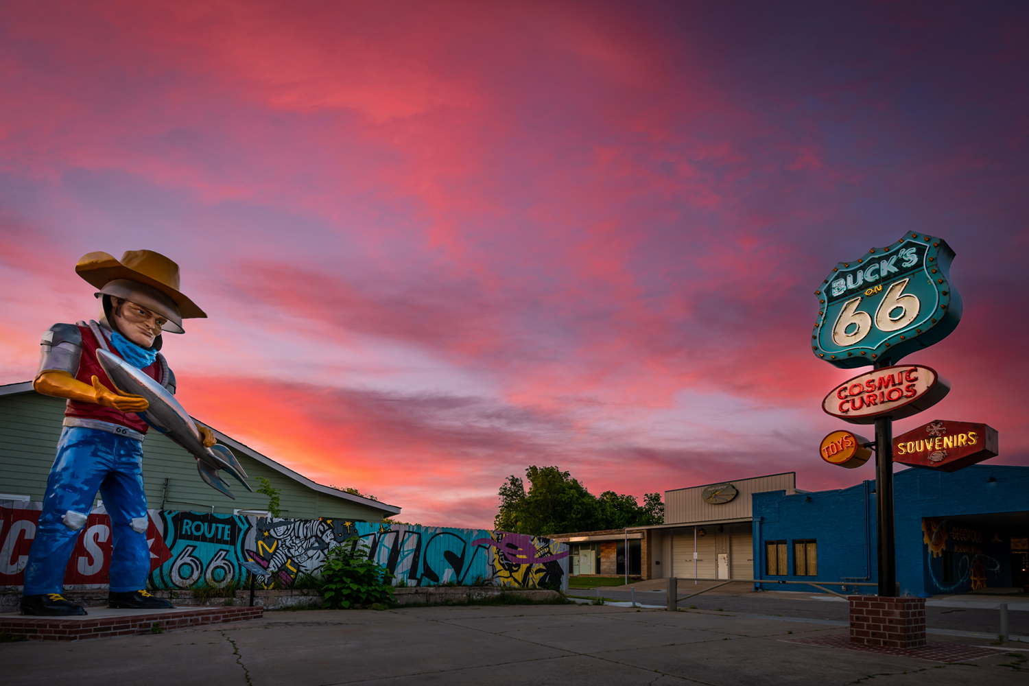 A giant statue of a cowboy holding a silver rocket outside a Route 66 shop with neon signs reading "Buck's on 66," "Cosmic Curios" and "Souvenirs," under a pink and purple sunset sky.