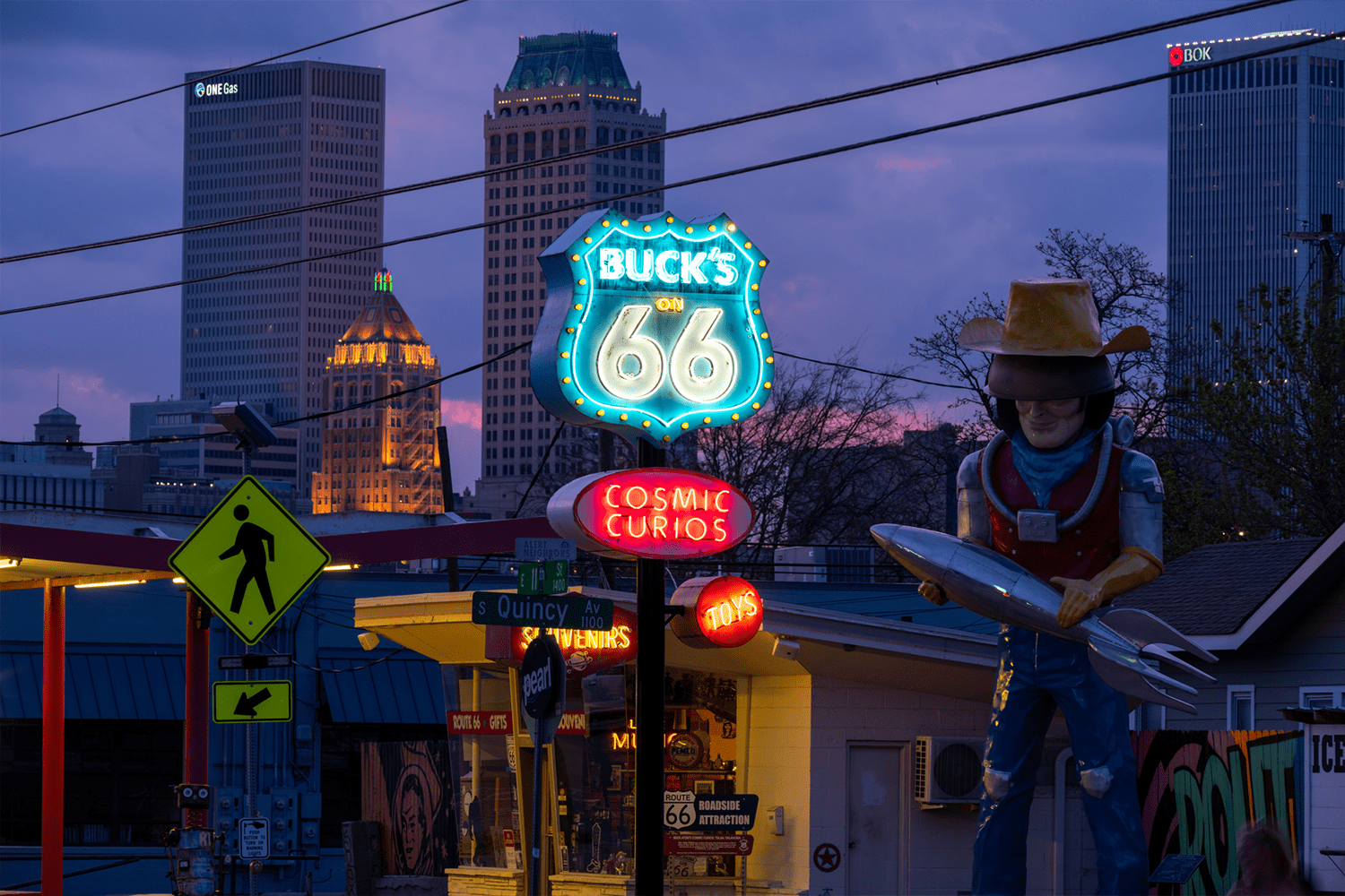 A neon sign reading "Buck's on 66" and "Cosmic Curios" beside a giant cowboy statue holding a rocket, with the downtown Tulsa skyline glowing at dusk in the background.