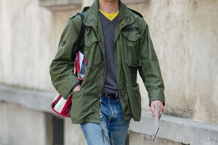 A man wearing dark yellow gray long sleeve sweater, dark green cargo jacket, navy blue ripped baggy denim jean pants, shiny black oxford moccasin leather shoes