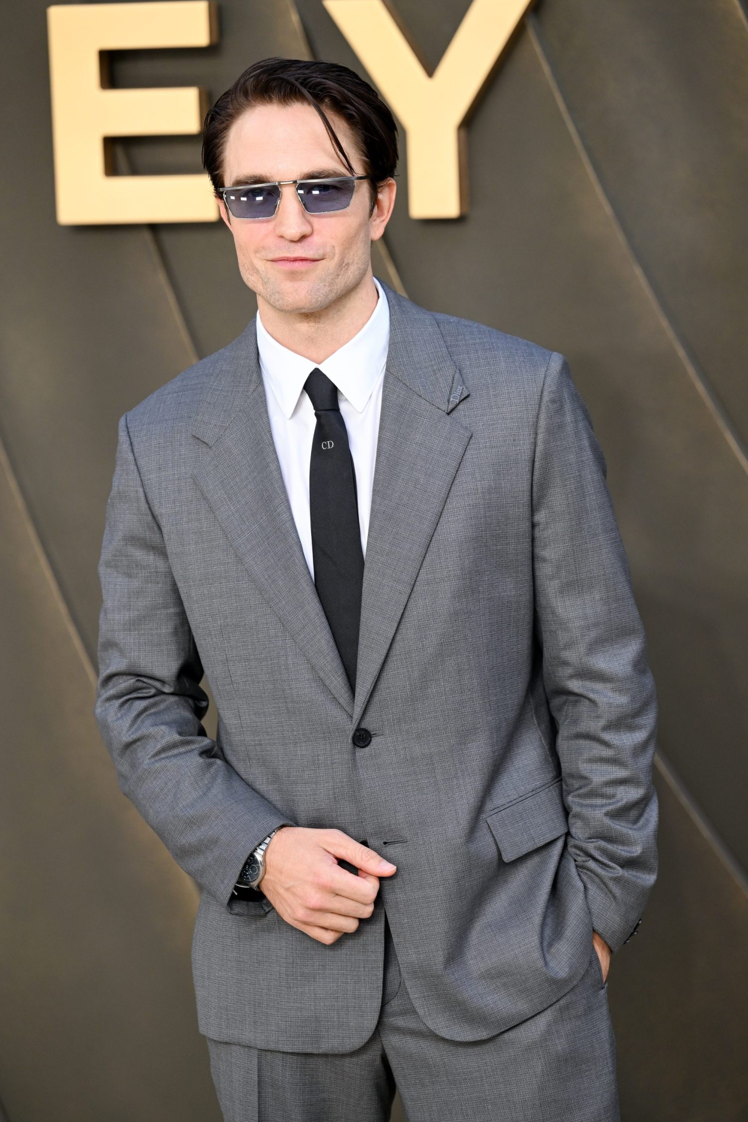 A man in a grey suit and sunglasses standing for a photo.