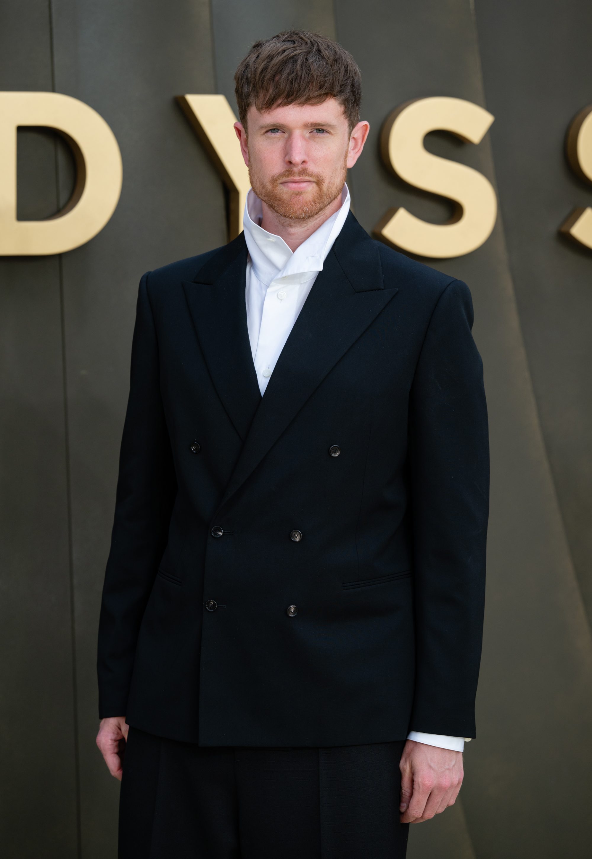 A man in a black suit and collared white shirt standing for a photo.