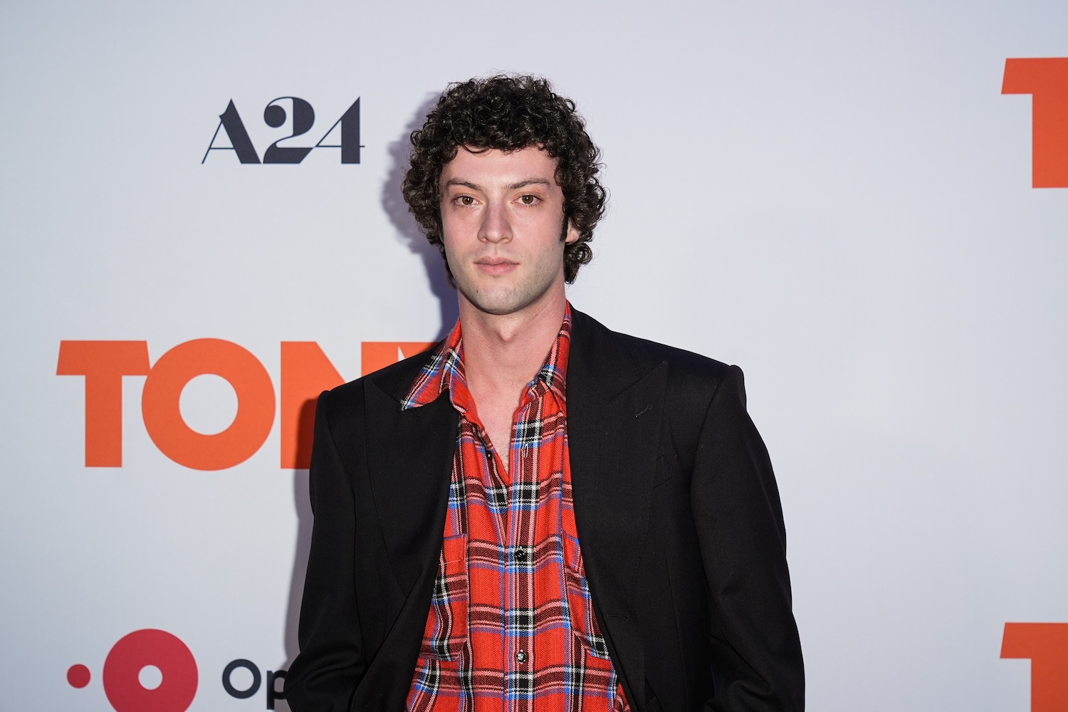 a man with curly black hair in a black suit and red plaid shirt