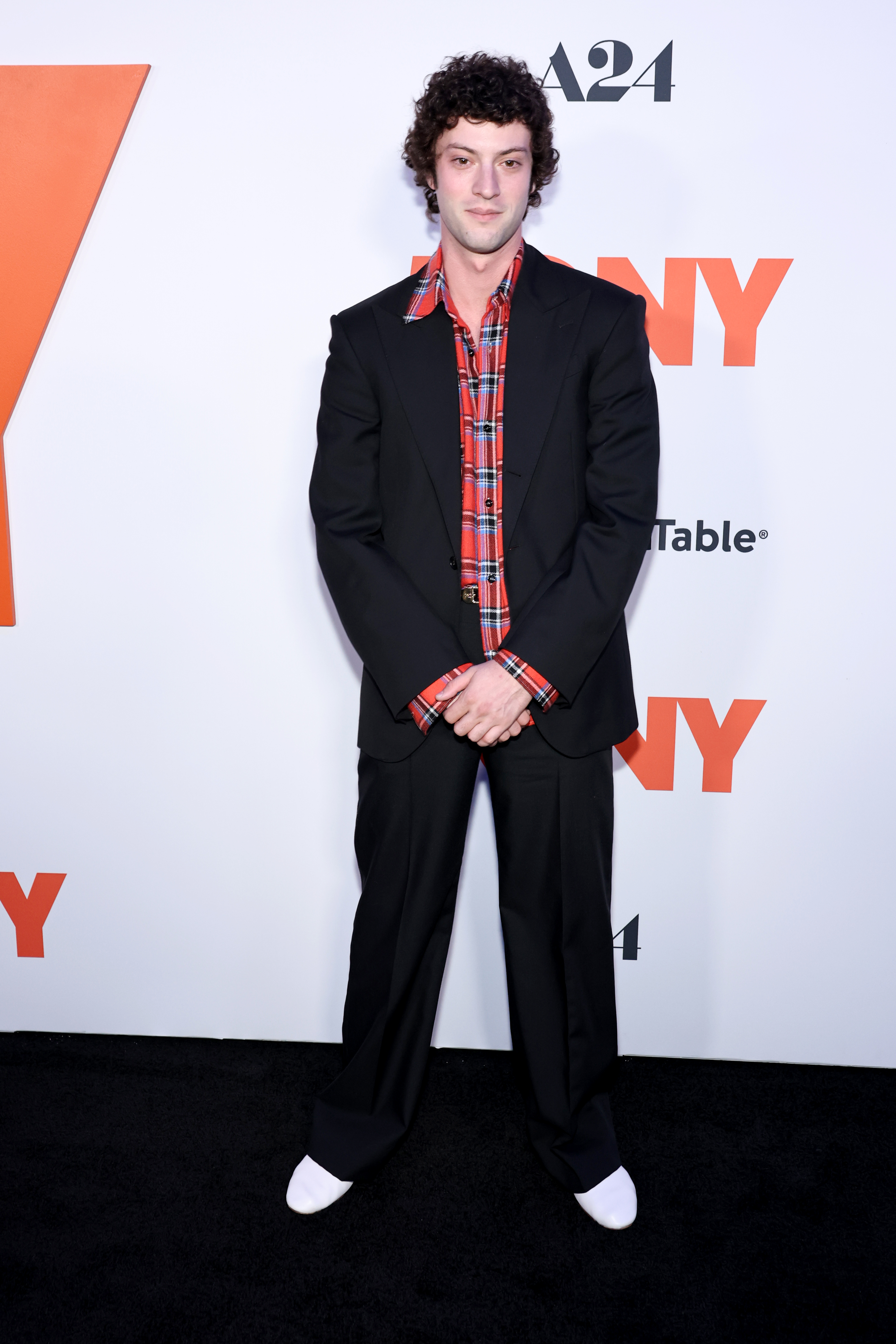 a man with curly black hair in a black suit and red plaid shirt