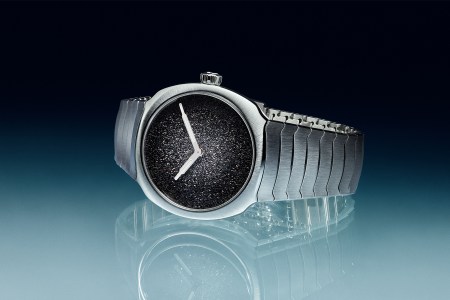 A stainless steel watch with two hands and a silver dial that fades from dark to light on a black and blue background
