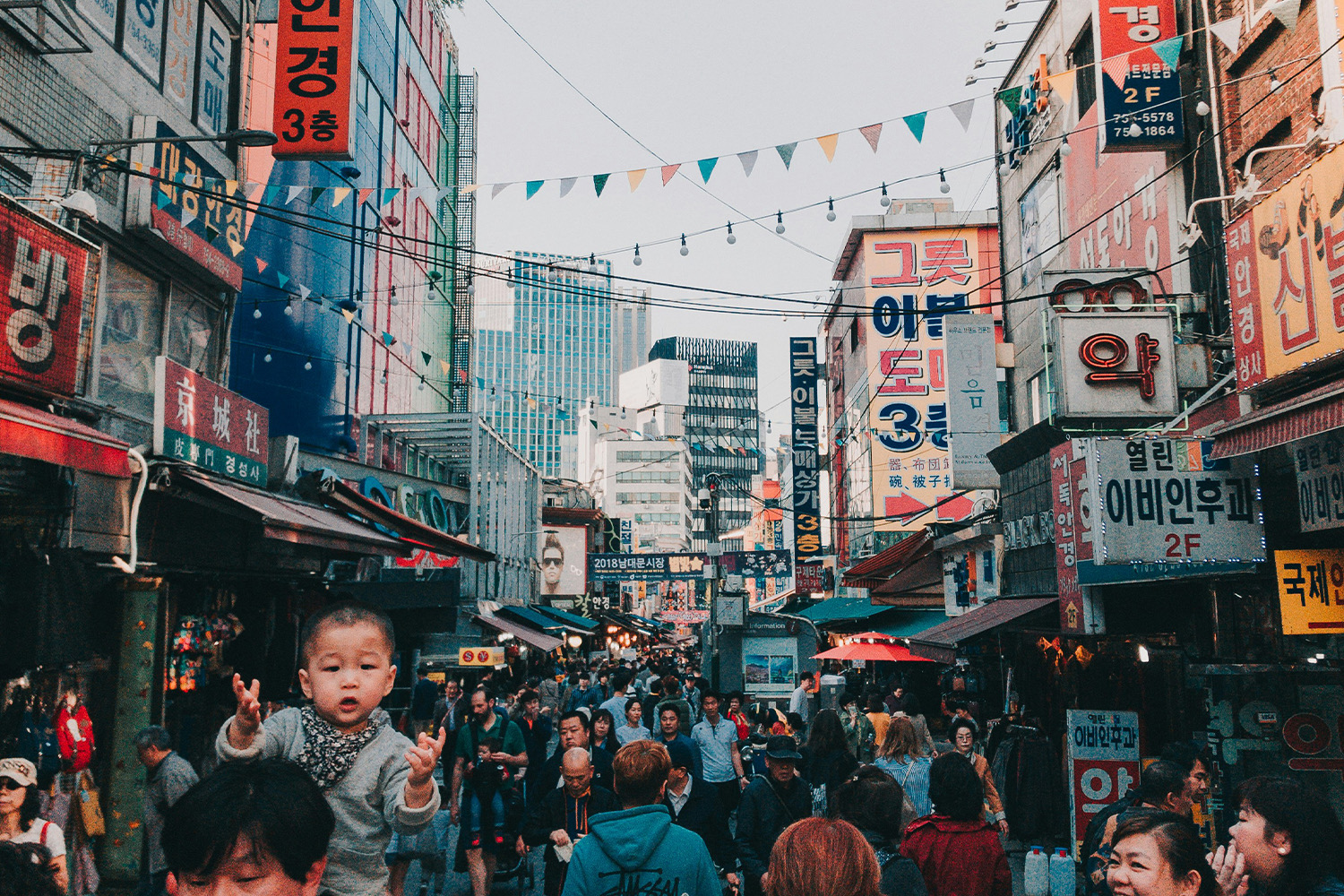 Bustling shopping street in Seoul filled with pedestrians beneath colorful Korean storefront signs and string lights, with high-rise buildings visible in the background, capturing the energy of the city's retail districts.