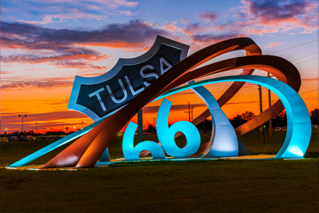 A glowing sculpture of a Route 66 shield reading "Tulsa" next to swooping metal ribbons shaped like "66," set against a fiery orange and purple sunset sky.