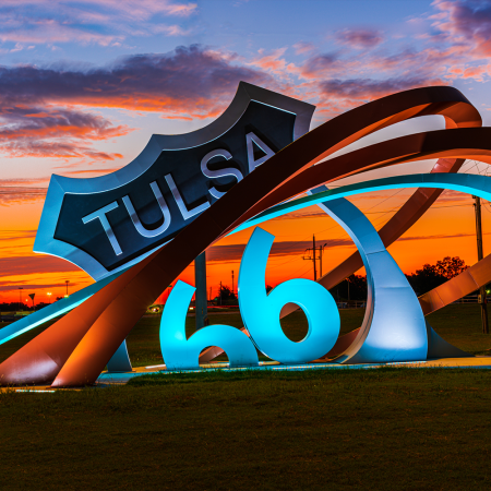 A glowing sculpture of a Route 66 shield reading "Tulsa" next to swooping metal ribbons shaped like "66," set against a fiery orange and purple sunset sky.