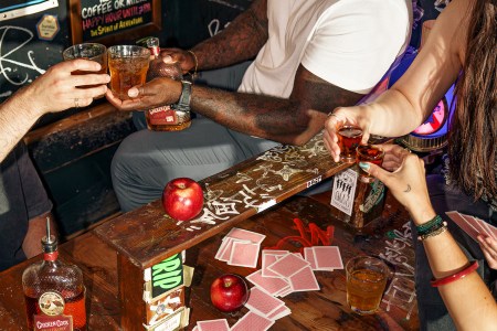 Several people's hands over a table holding glasses of whiskey, with a bottle of Chicken Cock Old Glory whiskey on the table.