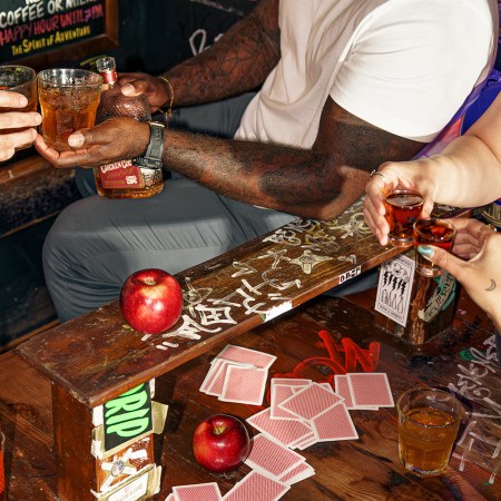 Several people's hands over a table holding glasses of whiskey, with a bottle of Chicken Cock Old Glory whiskey on the table.
