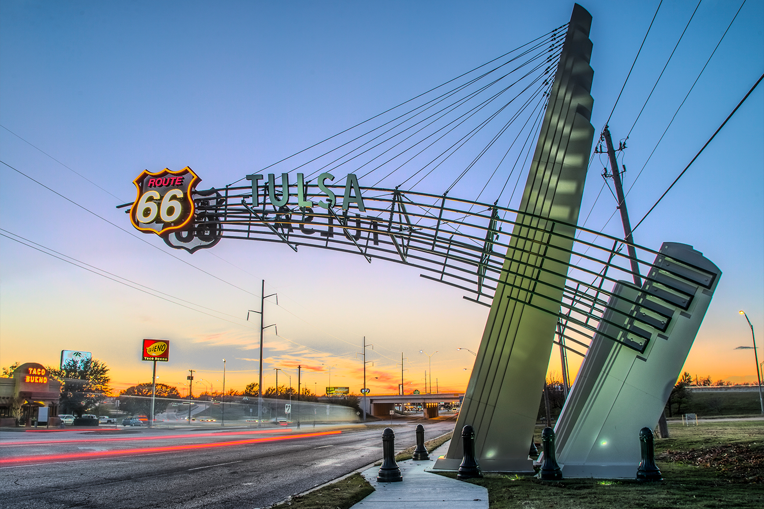 An illuminated cable-stayed arch reading "Tulsa" with a neon Route 66 shield below it, spanning a road at dusk with light trails from passing cars.
