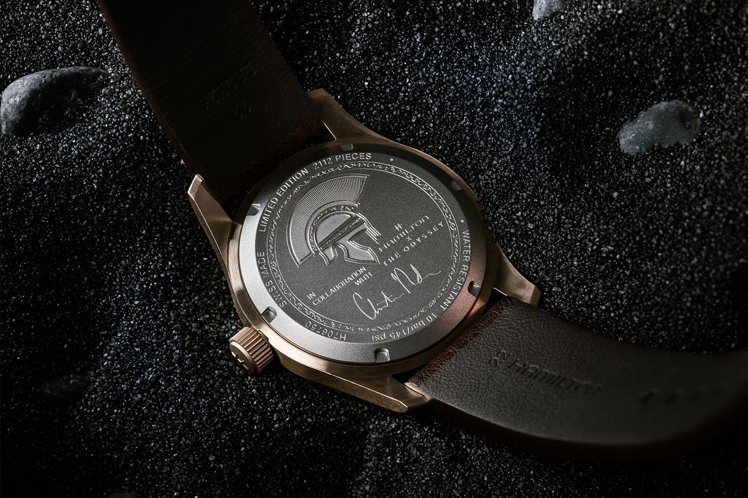 A watch lying on volcanic soil showing the titanium caseback, which is engraved with the helmet of Odysseus and the signature of Odyssey director Christopher Nolan