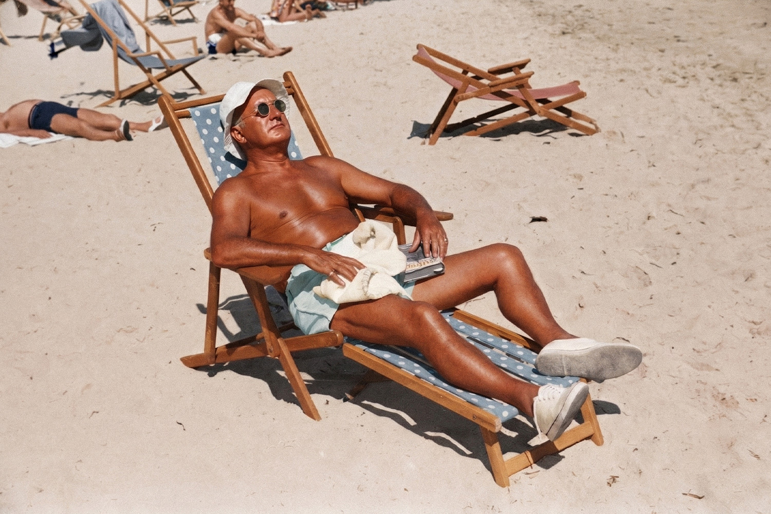 An old man relaxes shirtless at the beach with sunglasses and a beach shoes on while laying on a beach chair.