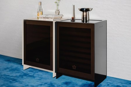 Two modern wine refrigerators, one white and one black, stand side-by-side on a blue carpet against a white brick wall.