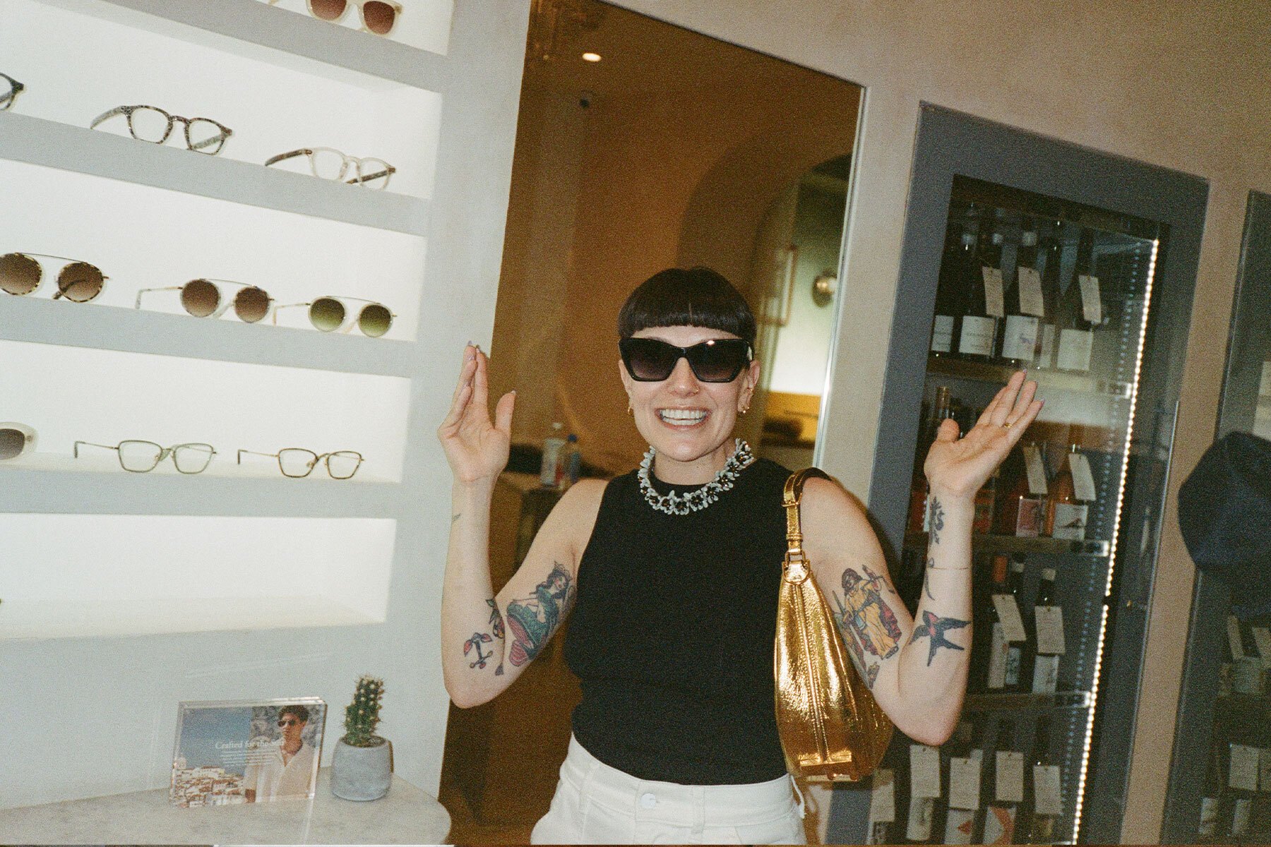 a woman in a black sleeveless top wearing dark sunglasses in a sunglasses store with a wine fridge in the background