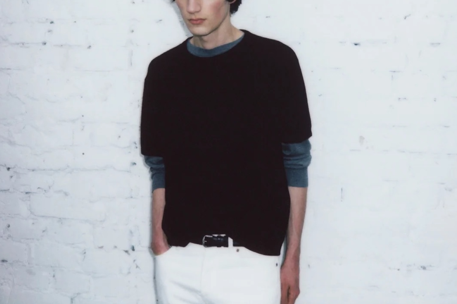 a model wearing a black shirt and white pants against a white concrete wall.