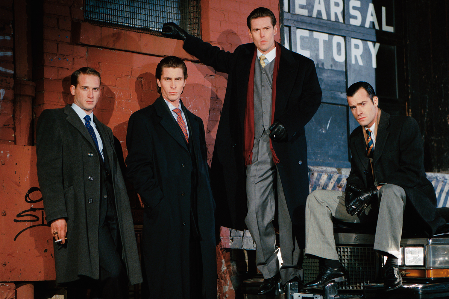 Four men in dark overcoats and suits pose against a brick wall and car, one holding a cigarette.