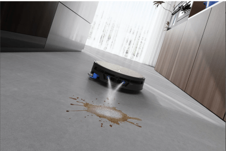 A black robotic vacuum cleaner spraying water onto a brown liquid spill on a gray floor in a modern kitchen.