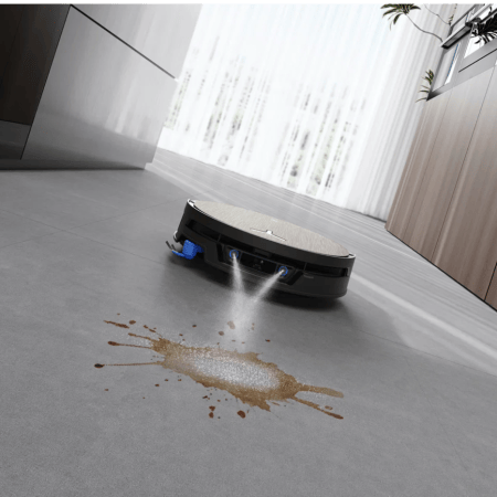 A black robotic vacuum cleaner spraying water onto a brown liquid spill on a gray floor in a modern kitchen.