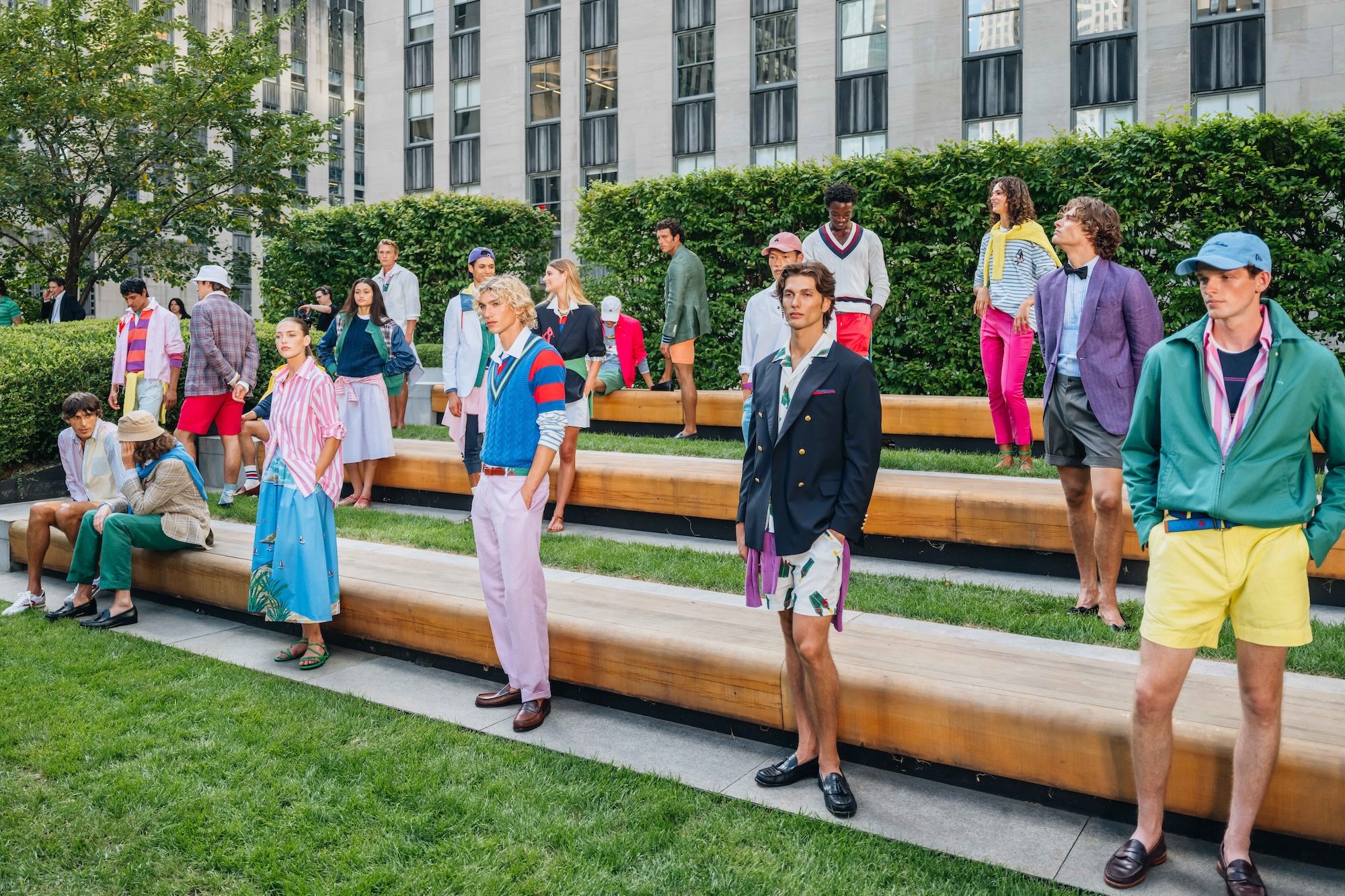 Models in colorful, preppy clothing pose on wooden benches in a garden with a city building backdrop.