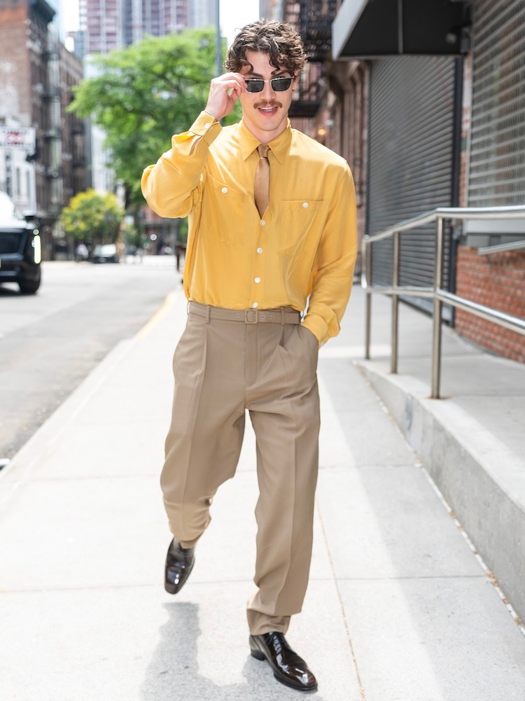 A man with curly hair and a mustache, wearing a yellow shirt, tan pants, and sunglasses, walks on a city sidewalk.