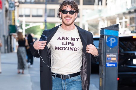 A smiling man in sunglasses and a black blazer over a t-shirt that reads "ARE MY LIPS MOVING?" holds a phone.