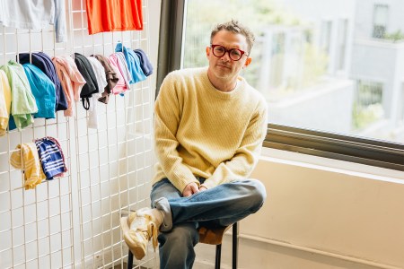 Man in yellow sweater and red glasses sits on a stool, with colorful clothes hanging on a grid behind him.