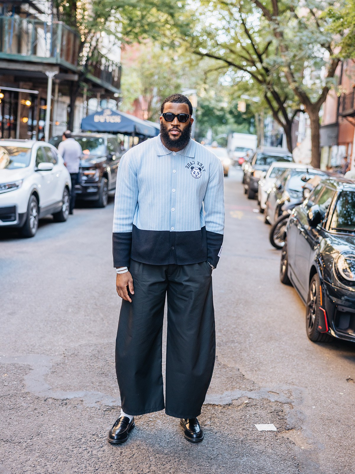 A man with a beard and sunglasses wears a light blue and black striped cardigan, wide black pants, and black loafers on a city street.