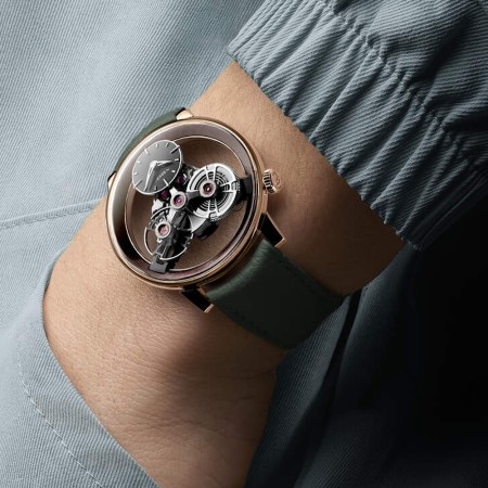 A person's arm wearing a rose gold watch with a clear face revealing intricate gears, on a dark green leather strap.