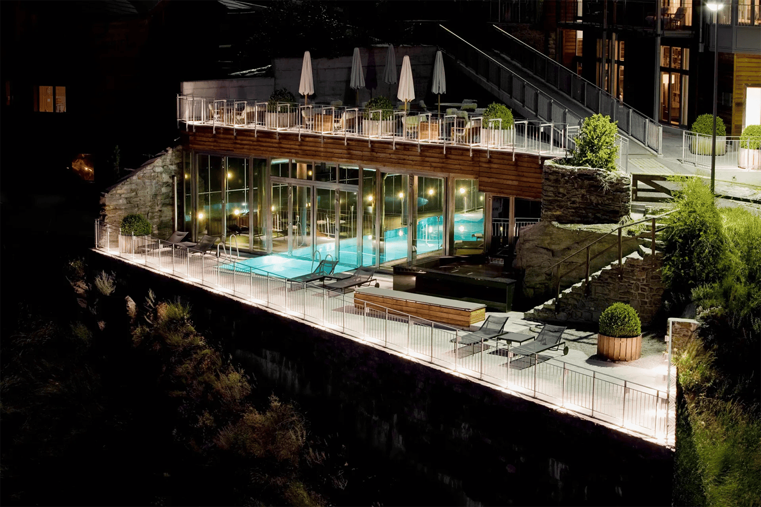 Illuminated outdoor pool and patio area with lounge chairs and umbrellas, next to a building with large glass windows, at night.