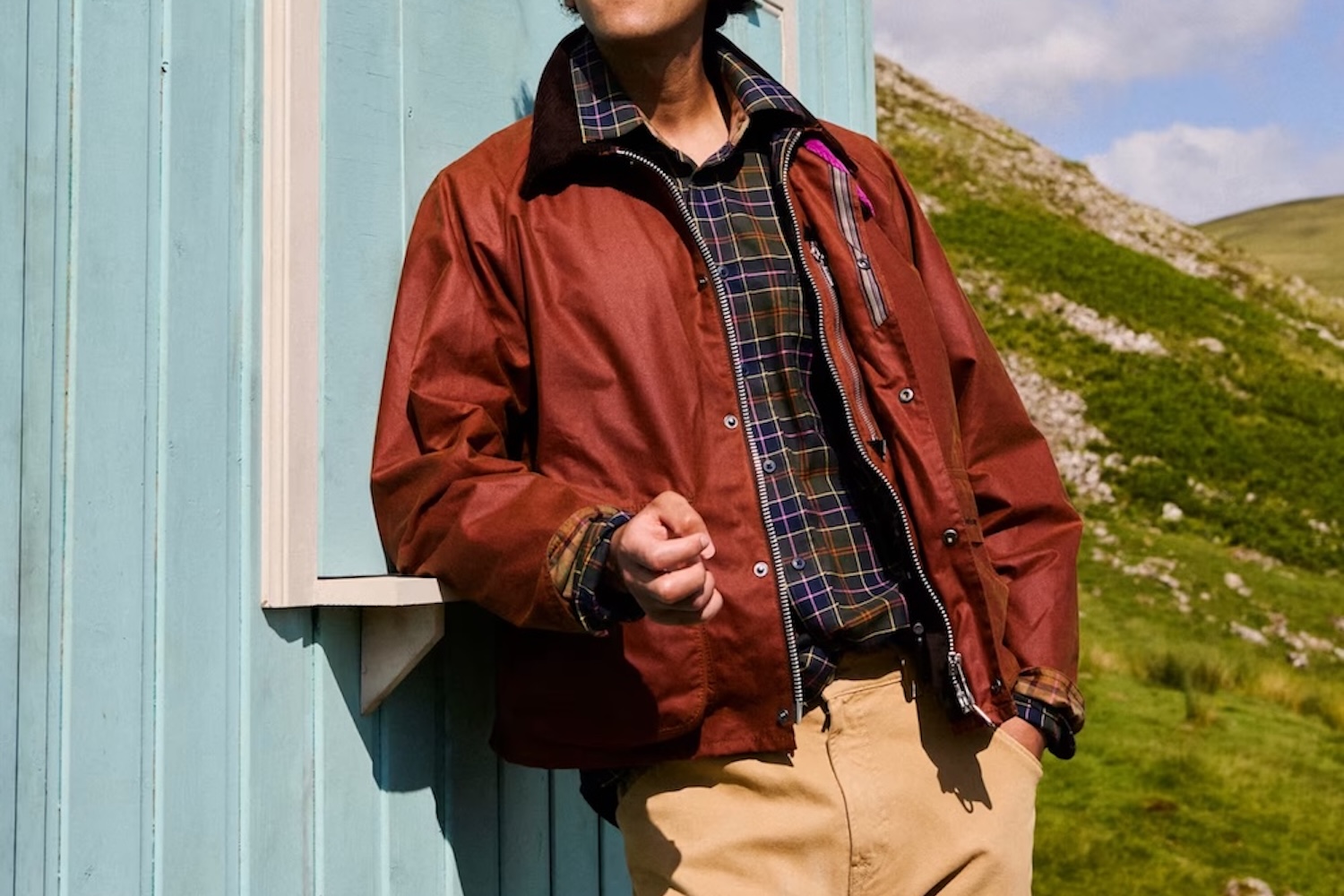 Person in a brown waxed jacket, plaid shirt, and tan pants leaning on a light blue wall, with a green hillside in the background.