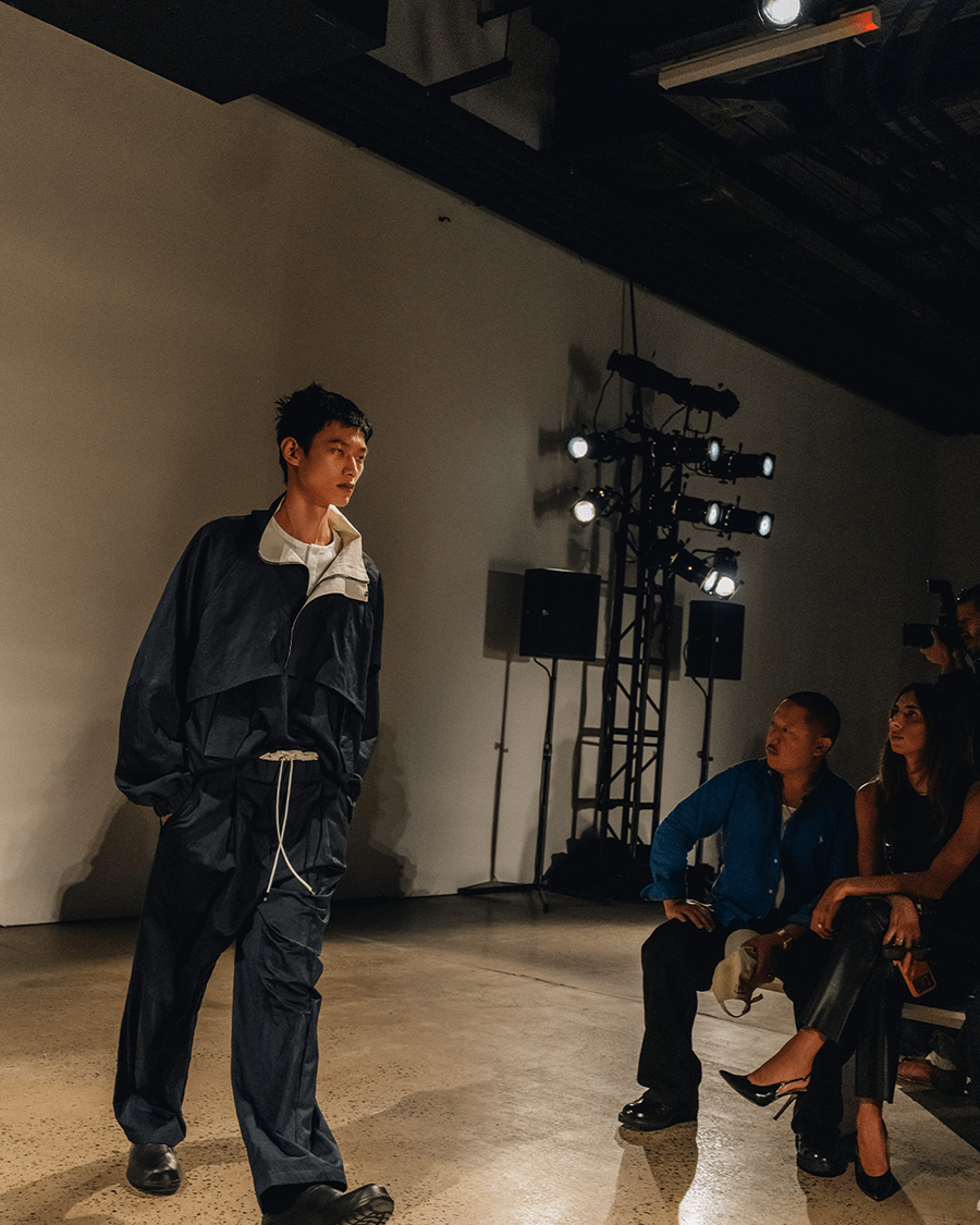 A male model in a dark blue tracksuit walks a runway, with audience members and stage lights in the background.