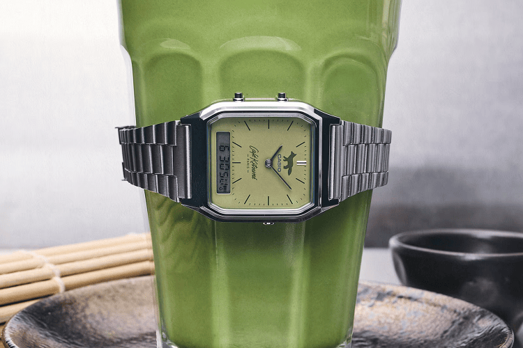 Silver Casio watch with a green face and fox logo, wrapped around a glass of green matcha tea.