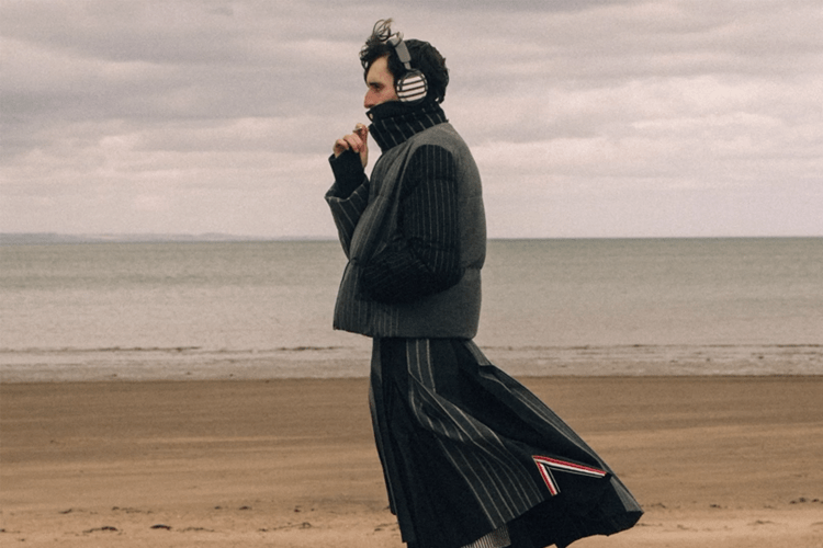 Man in a striped puffer vest, turtleneck, and pleated skirt with headphones on a sandy beach.