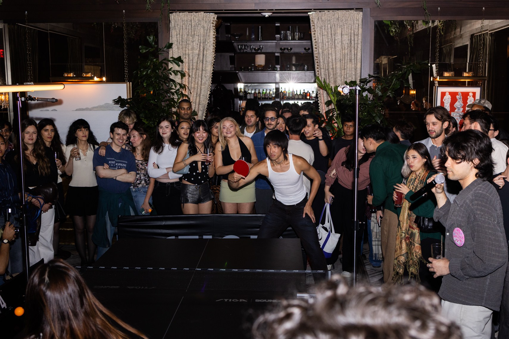 A group of young adults gathered around a ping-pong table, one man playing while another sings into a microphone.