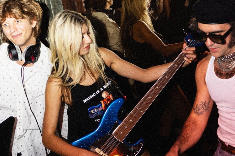 A blonde woman in a "Music is a Blessing" tank top plays a blue glitter bass guitar, flanked by a smiling DJ and a man in a beret.