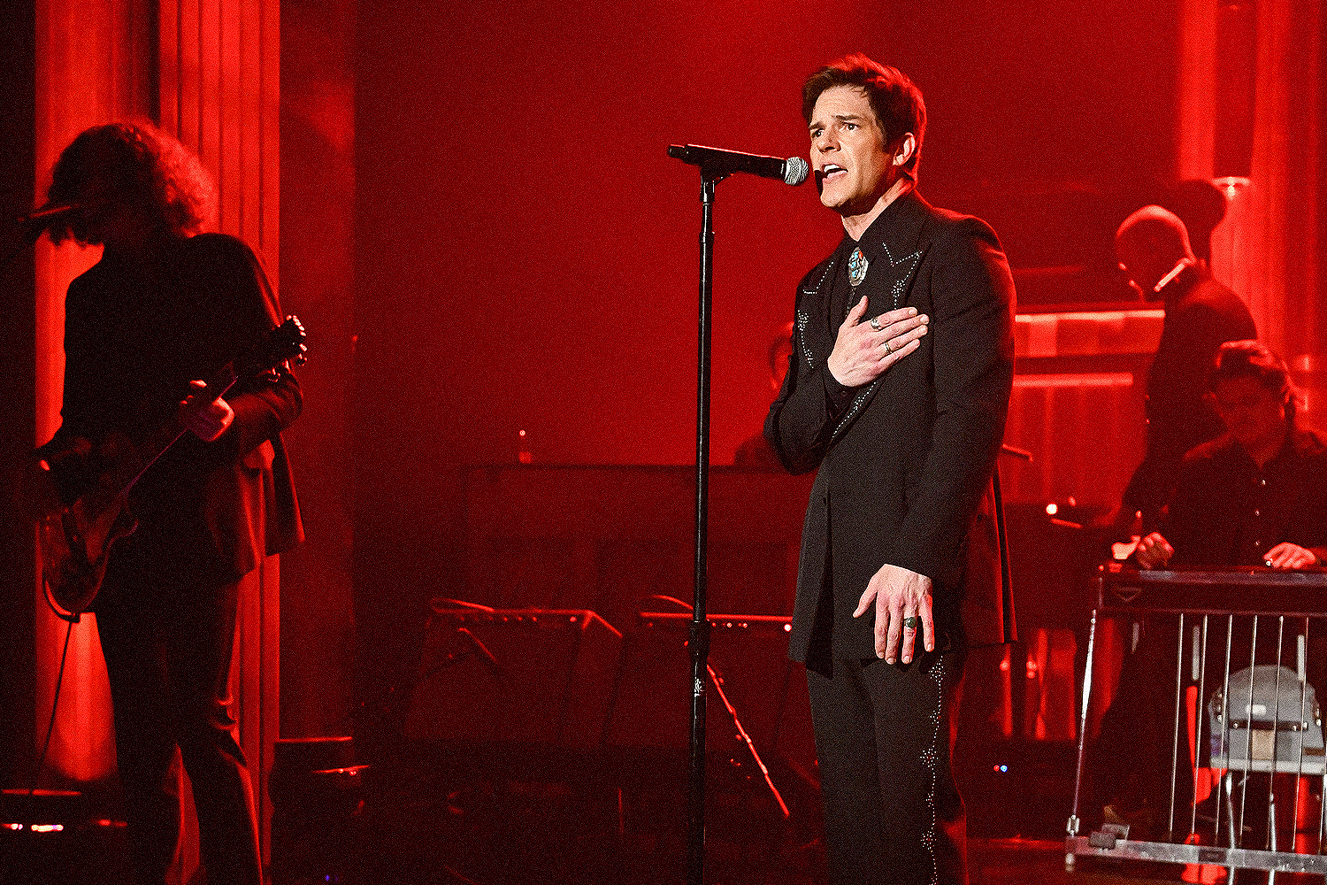 Singer Brandon Flowers in a black suit with a bolo tie and rings, hand on his chest, singing into a microphone on a red-lit stage