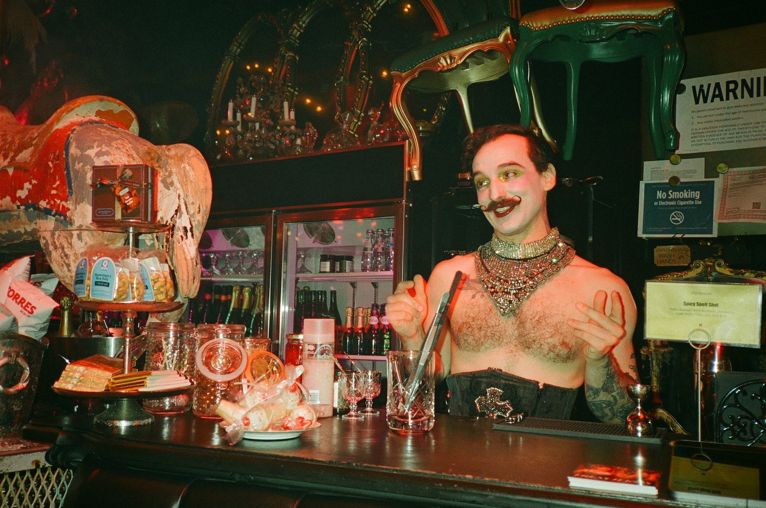 A smiling, shirtless person with a handlebar mustache, glittery makeup, and a large jeweled necklace behind a bar.
