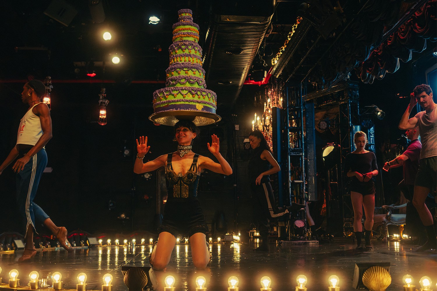 A performer kneels on stage, balancing a large, tiered cake on her head, surrounded by other performers and stage lights.