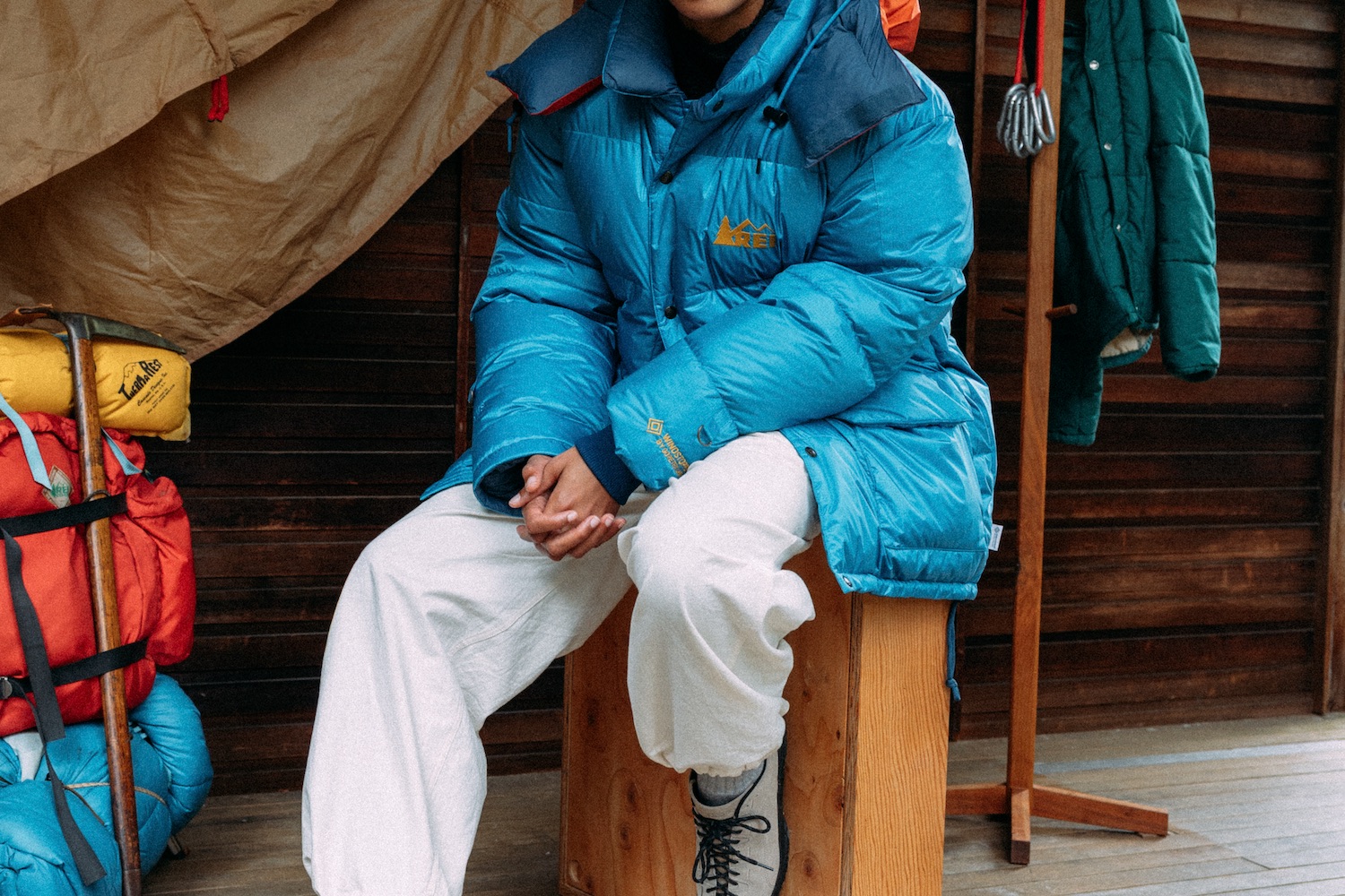 Person in blue puffer jacket and white pants sits on a wooden box, with camping gear nearby.