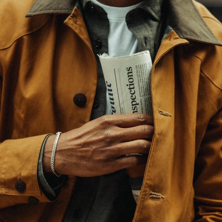 A person in a mustard yellow jacket with a dark collar holds a folded newspaper in their left hand.